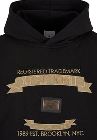 Sweat à capuche noir avec texte brodé beige "Registered Trademark Karl Kani", plaque métallique de marque et "1989 Est. Brooklyn, NYC" sur le devant.