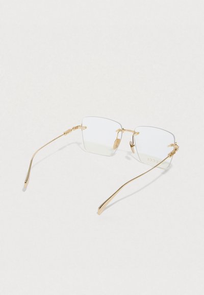 Négyzet alakú szemüveg vékony aranyszínű fémkerettel és átlátszó lencsékkel. A szárakon díszítő elemek találhatók, és letisztult, minimalista kialakítású.