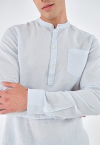 Lichtblauw, langemouwhemd van linnen, met een opstaande kraag, een halve knoopsluiting en een borstzak. Gladde textuur.