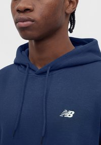 Jong persoon met gevlochten haar die een donkerblauwe New Balance hoodie draagt, waarbij de bovenste borst en het gedeeltelijke gezicht tegen een effen achtergrond te zien zijn.