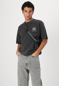 Černé oversized tričko s grafickým potiskem na hrudi, spárované se světle šedými džíny; model má na sobě crossbody tašku.