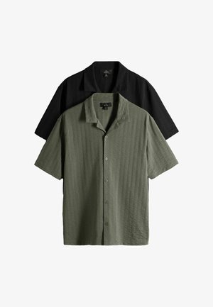 Uomo con capelli ricci che indossa una camicia oliva a maniche corte con fantasia, pantaloni neri e scarpe beige, in piedi con le mani nelle tasche guardando verso il basso.
