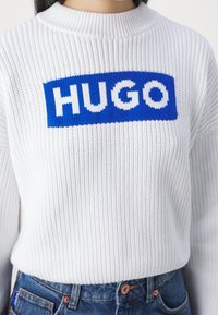 Maglione bianco lavorato a maglia con una texture a coste, caratterizzato da un logo rettangolare blu con la scritta "HUGO" in lettere bianche sulla parte anteriore.