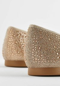 Gros plan sur des chaussures plates beiges ornées de nombreux petits strass brillants couvrant la tige en tissu et les semelles en caoutchouc marron.