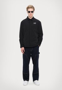 Svart lättvikts zip-jacka med krage och EA7-logotyp, kombinerad med marinblå löst sittande byxor med röda sömnadsdetaljer och fickor.