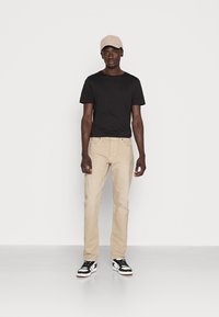 Calças de algodão bege, t-shirt preta e boné cinza claro, usados com ténis preto e branco. Modelo a posicionar-se contra um fundo simples.