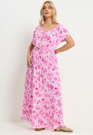 Kvinde iført en hvid maxikjole med pink blomsterprint, korte ærmer og taljefasong, stående med den ene hånd på hoften og iført sandaler.