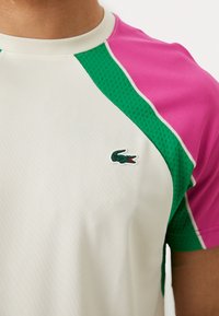 Light beige athletic shirt with green and pink accents, featuring a textured finish and a small green crocodile logo on the chest.