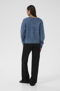 Vastag, kék kötött pulóver bordás alsó résszel és oversize ujjal, fekete, bő szabású nadrággal és lapos cipővel párosítva. Egyszerű, texturált dizájn.