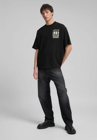 T-shirt oversize nera con stampa grafica sul petto, abbinata a jeans scuri e stivali neri chunky, su uno sfondo neutro.