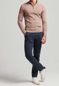 Jasnobrązowy sweter z zamkiem na szyi i długimi rękawami, w połączeniu z ciemnoniebieskimi chinosami i białymi trampkami. Prosty design, bez wzorów i ozdób.