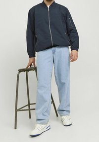 Marinblå bomberjacka med dragkedja framtill och sidofickor, kombinerad med ljusblå jeans och vita sneakers på en metallpall.