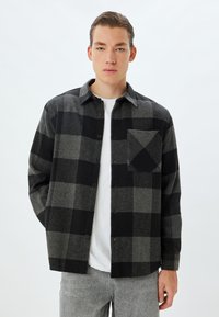 Czarna i szara koszula w kratę z wełny, z zapięciem na guziki, kołnierzykiem oraz pojedynczą kieszenią na piersi z ukośnym wzorem.