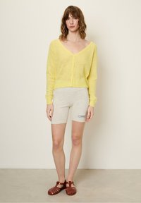 Gelbe leichte Strickjacke mit tiefem V-Ausschnitt und Knopfverschluss; kombiniert mit grauen Shorts, die ein Etikett haben, und braunen Riemchensandalen.