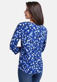 Langärmliges blaues Oberteil mit einem unregelmäßigen Muster aus hellblauen, weißen und schwarzen Formen. Der Rücken hat einen abgerundeten Saum und eine weiche Textur.