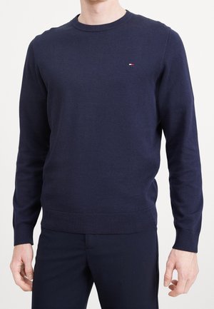 Homme portant un pull manche longue bleu marine avec un petit logo rouge, blanc et bleu sur le côté gauche de la poitrine.