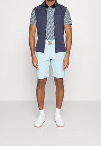 Navy ärmlös väst över en mönstrad blå pikétröja, kombinerad med ljusblå shorts, vitt bälte och vita golfskor. Slät, avslappnad design.