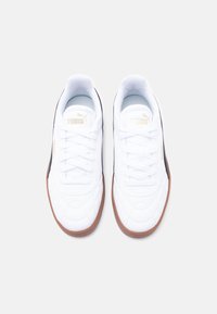 Ζευγάρι λευκά αθλητικά παπούτσια Puma με καφέ σόλες από καουτσούκ, σχεδιασμό επένδυσης στο πάνω μέρος και χρυσά λογότυπα Puma στις γλώσσες και τις πλευρές.