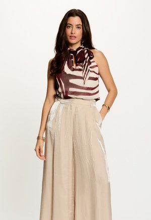 Mujer con el pelo largo y oscuro que lleva un top sin mangas burdeos y blanco con estampado y lazo, y pantalones beige de pierna ancha y talle alto.
