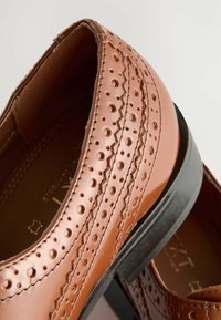 Brown leather loafers with perforated detailing and zigzag stitching, featuring a flat black rubber sole and smooth inner lining.