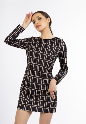 Frau trägt ein langärmliges Mini-Kleid mit schwarz-beigem geometrischem Muster und Knopfdetails an Manschetten und Schultern, posiert mit der Hand nahe am Kopf.
