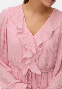 Rosa plisserad blus med V-hals, volangkrage och långa ärmar. Har ett elastiskt midjeband med en knytdetalj. Mjuk, texturerad tyg.