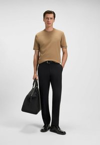 Short-sleeve tan shirt with white trim, black trousers, and black dress shoes. Carrying a black duffel bag, standing against a plain background.