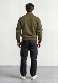 Giacca bomber verde oliva con collo alto, polsini e vita a costine. Abbinata a pantaloni neri e calzature bianche. Texture liscia.