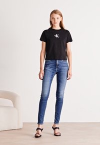 Zwarte katoenen t-shirt met een logo, gecombineerd met blauwe skinny jeans. Model draagt zwarte hoge hakken. Eenvoudig, getailleerd ontwerp, minimale details.