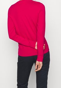 Maglione aderente a maniche lunghe di un vivace rosa con polsini e fondo a costine. Indossato con pantaloni scuri, che mettono in risalto una texture liscia.