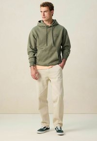 Man staat met handen in de zakken, gekleed in een olijfgroene hoodie, beige broek, perzikkleurig t-shirt en zwart-witte sneakers tegen een beige achtergrond.
