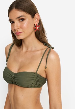 Mlada žena u maslinastozelenom bikinku s vezicama i velikim naušnicama u obliku crvenih zvijezda morskih zvijezda, pozira na bijeloj pozadini.