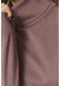 Gros plan d'une personne portant un pull à col roulé côtelé mauve, tenant le col près du cou avec la main gauche.