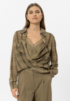 Woman with long brown hair wears green lace top under a brown plaid button-up shirt and matching high-waisted pants with one hand in pocket.