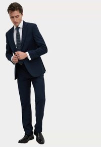 Completo blu scuro con vestibilità slim, in tessuto liscio; dotato di giacca a un bottone, camicia bianca e cravatta a righe; scarpe nere eleganti.