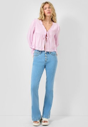Femme aux longs cheveux blonds portant un chemisier rose clair noué, un jean taille haute bleu clair avec boutons, et des sandales blanches à plateforme.