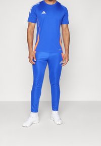 T-shirt et pantalon de sport bleus en tissu lisse et extensible, avec des accents orange et des rayures blanches, associés à des chaussures de sport blanches.