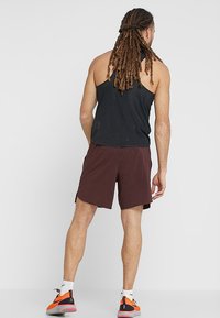 Hombre con rastas que lleva una camiseta sin mangas negra, pantalones cortos color burdeos, calcetines blancos y zapatillas de correr naranjas, de pie de espaldas.