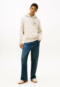Sudadera color beige claro con un bolsillo frontal, capucha con cordones y un pequeño logo; combinada con unos jeans de denim azul holgados y zapatos de color claro.