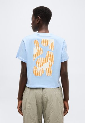 Osoba s krátkymi vlasmi, ktorá má na sebe svetlomodré krátke tričko s oranžovo-bielym abstraktným vzorom na chrbte a béžové nohavice.