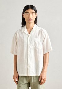 White short-sleeve shirt with a textured pattern, featuring a chest pocket and six buttons down the front. Lightweight, casual fit.