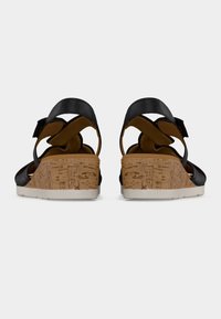 Paire de sandales compensées en cuir noir avec talons en liège et semelles blanches, vues de dos sur un fond clair uni.