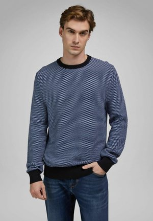 Jeune homme aux cheveux châtain clair portant un pull bleu à motifs avec des poignets noirs et un jean bleu, debout devant un fond gris.