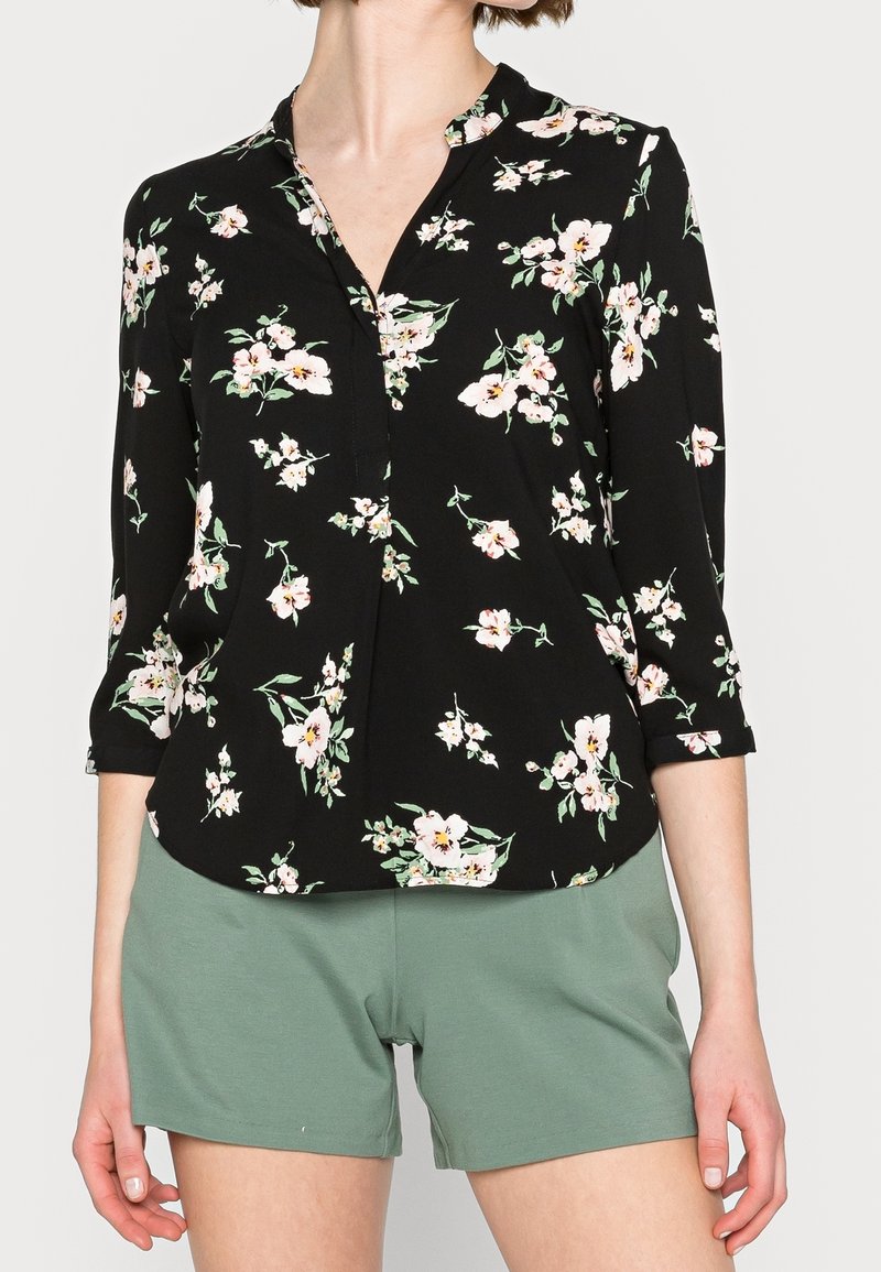 Donna che indossa una blusa nera a fiori con maniche a tre quarti e pantaloni corti verde chiaro, in piedi su uno sfondo semplice.