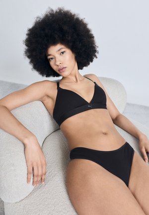 Žena s afro účesom sa nakláňa na textúrovanom béžovom kresle, má na sebe čiernu podprsenku a zodpovedajúce čierne spodné prádlo.