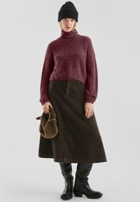 Burgundowy, puszysty sweter z wysokim kołnierzem, zielona spódnica z kordurowej tkaniny, czarne kozaki do kolan i brązowa torebka futrzana. Proste, teksturowane materiały.