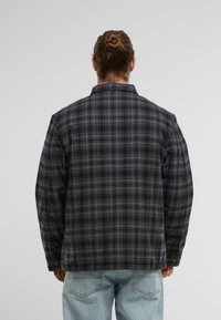 Melnā un pelēkā svītraina krekls ar brīvu piegriezumu, pogām aiztaisāmām piedurknēm un mīkstu tekstūru. Redzams piešuvums un standarta apkakle.