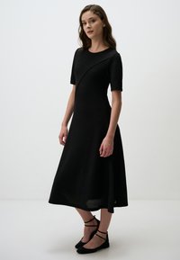 Robe en maille noire avec un corsage ajusté et une jupe évasée, présentant des manches courtes et un détail de design asymétrique. Accompagnée de ballerines noires.
