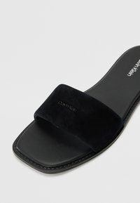 Svart glidesandal med en myk semsket stropp og en flat, rektangulær såle. Har en sømdetalj langs kanten og et preget logo.