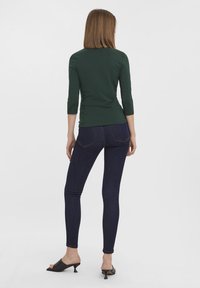 Un top ajustat cu mânecă trei sferturi de culoare verde închis, asortat cu blugi skinny de culoare albastră închis și mule negre cu vârful deschis, văzut din spate.